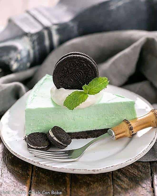Minty, green, frozen No-Bake Dessert with an Oreo Crust - Mint lovers will swoon with every bite of this frozen no bake dessert! Slice of frozen Pie with an Oreo Crust on a white dessert plate with Oreos and a bamboo handled fork.