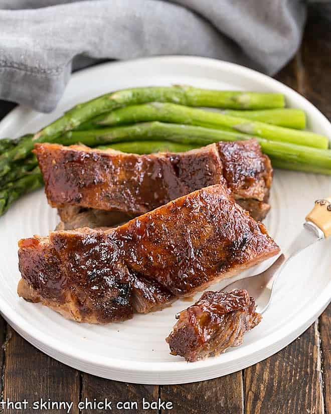 Tender bite of rib meat on a fork removed from a plate with two ribs and asparagus.