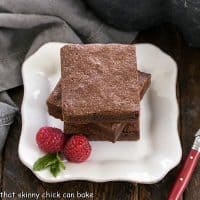3 fudgy brownies stacked on a square white plate with 2 raspberries and a sprig of mint