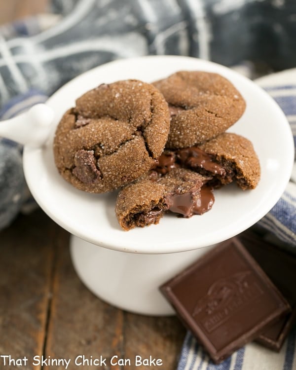 Chocolate Gingerbread Cookies on a small white pedestal, with one broken in half to reveal the melty chocolate interior