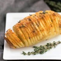 Hasselback potato on a white plate with a sprig of thyme