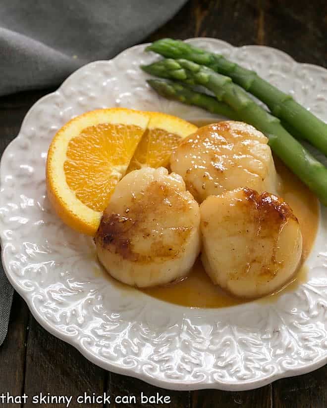 Pan seared scallops on a decorative white plate with orange slices and asparagus.