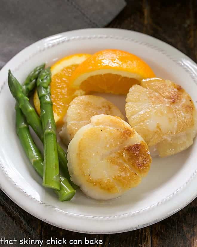 Scallops, asparagus and orange wedge on a white ceramic plate.