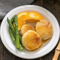Overhead view of pan seared scallops on a white plate with asparagus and fresh orange slices