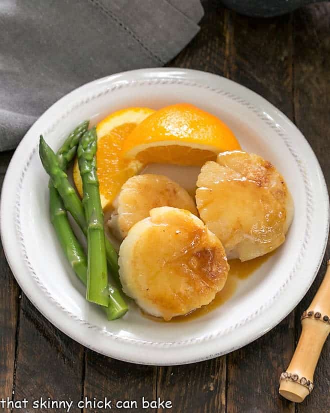 Overhead view of pan seared scallops on a white plate with asparagus and fresh orange slices