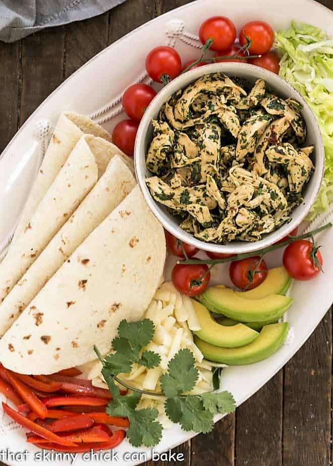 Serving platter with bowl of shredded chicken, tortillas and toppings with a cilantro garnish.