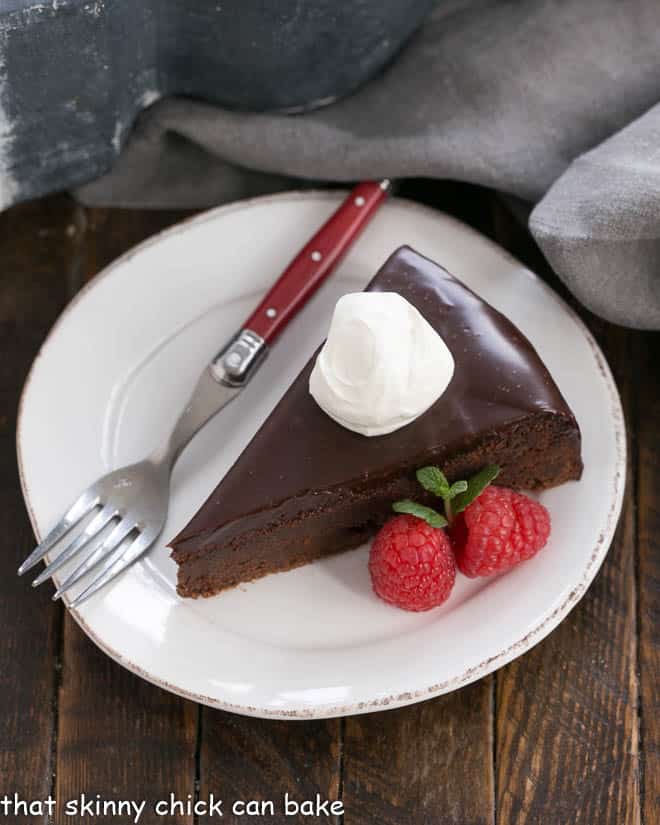 Overhead view of a slice of Flourless Chocolate Cake with Ganache Topping on a white plate with a red handled fork.