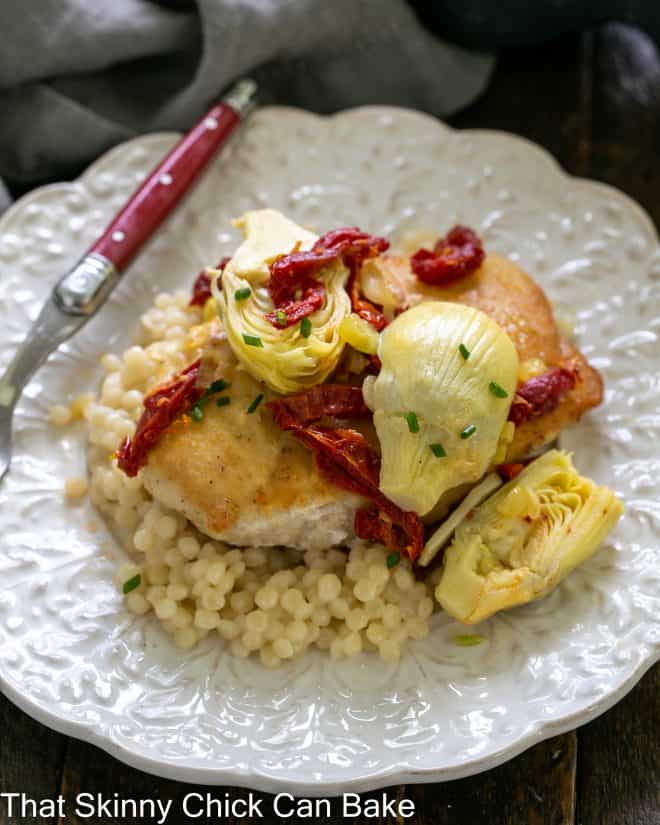 close overhead view of chicken with artichokes and sundried tomatoes over couscous on a white plate with a red handled fork