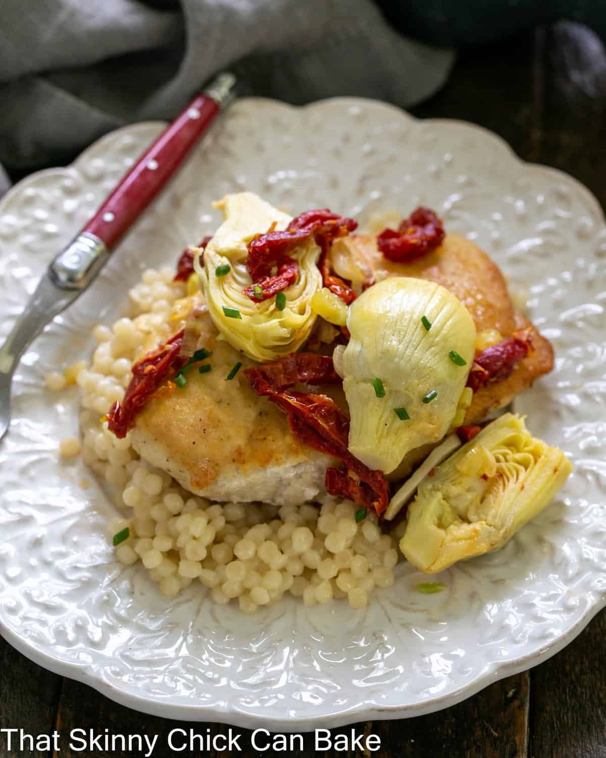 close overhead view of chicken with artichokes and sundried tomatoes over couscous on a white plate with a red handled fork.
