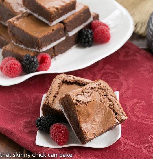 One Bowl Fudgy Brownies stacked on a serving tray with a small white plate with two brownies and berries in the foreground.