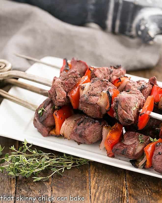 Marinated Greek Lamb Kebabs - Mediterranean flavors and a stint on the grill make for an out of this world entree! Marinated Greek Lamb Kebabs on a white serving tray with thyme garnish