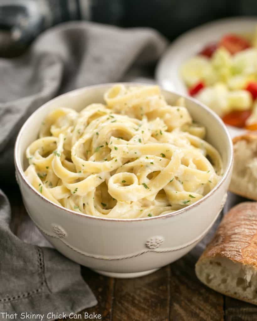 Light Fettuccine Alfredo in a white bowl with bread and salad in the back ground.