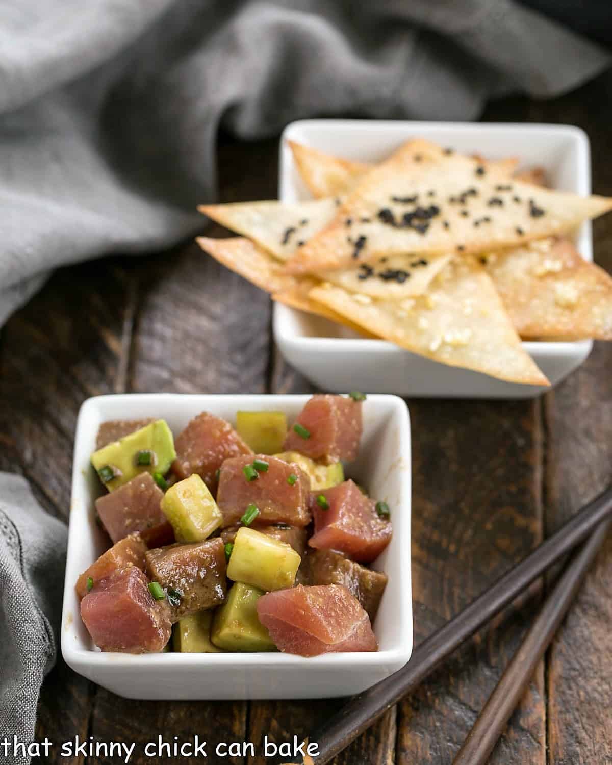 A square bowl of tuna tartare in front of wonton crisps in small white bowl.