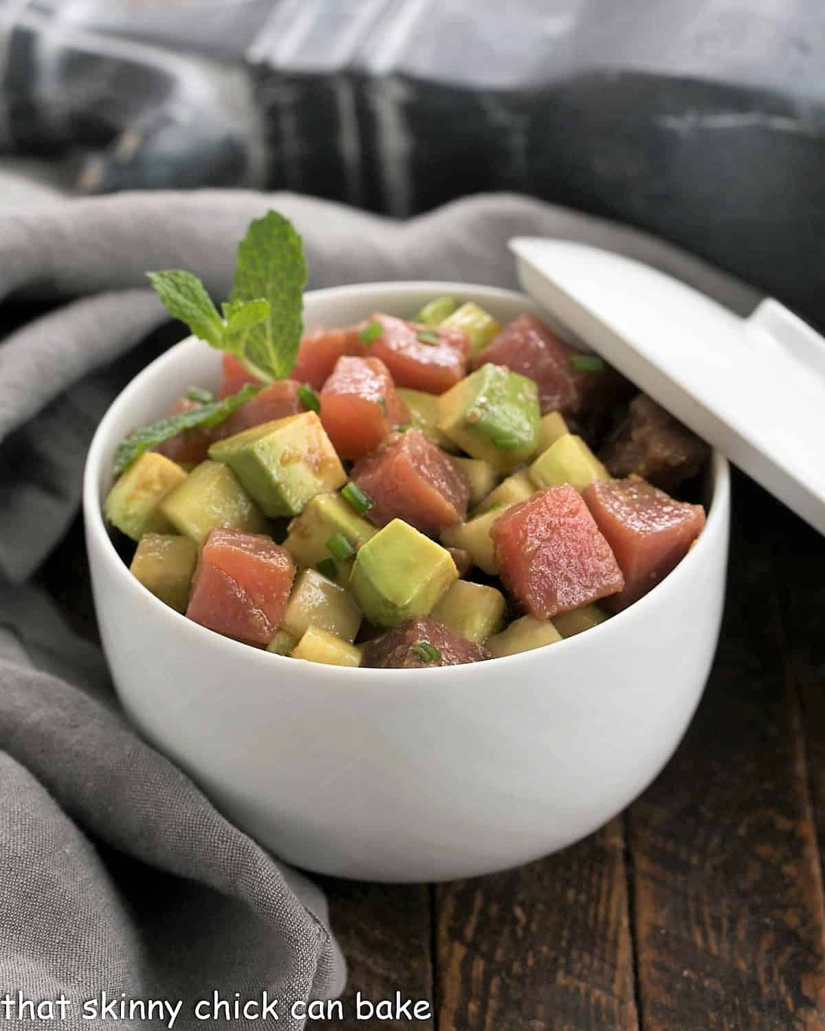 Tuna Tartare with Avocados in a white ceramic bowl with a lid propped on the side.