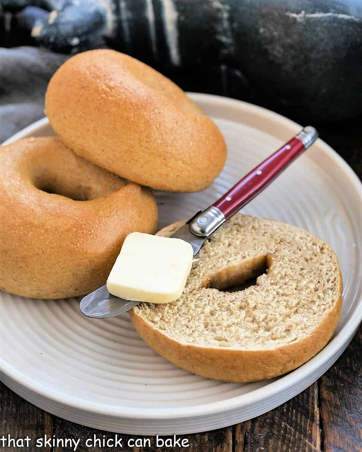 2 whole wheat bagels on a large white plate with one cut bagel topped with a red handle knife with a pat of butter.