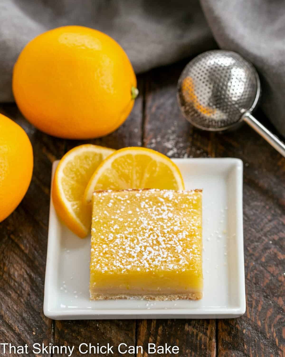 A Meyer Lemon bar on a white ceramic plate with lemon slices to garnish and a wand for powdered sugar.