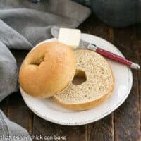 One Sliced whole wheat bagel on a white plate with a red handle knife topped with a pat of butter