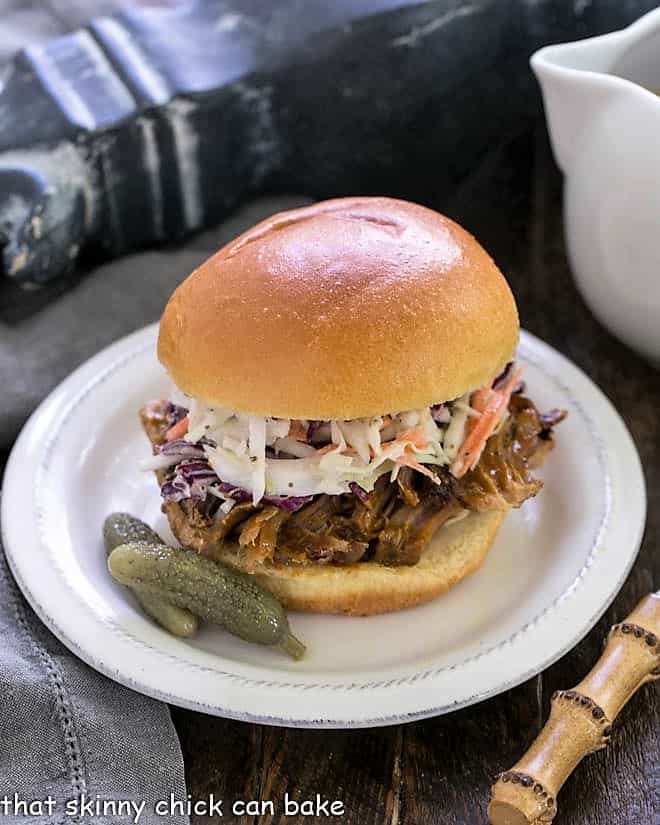 Best Pulled Pork Recipe topped with coleslaw on a white plate with pickles.