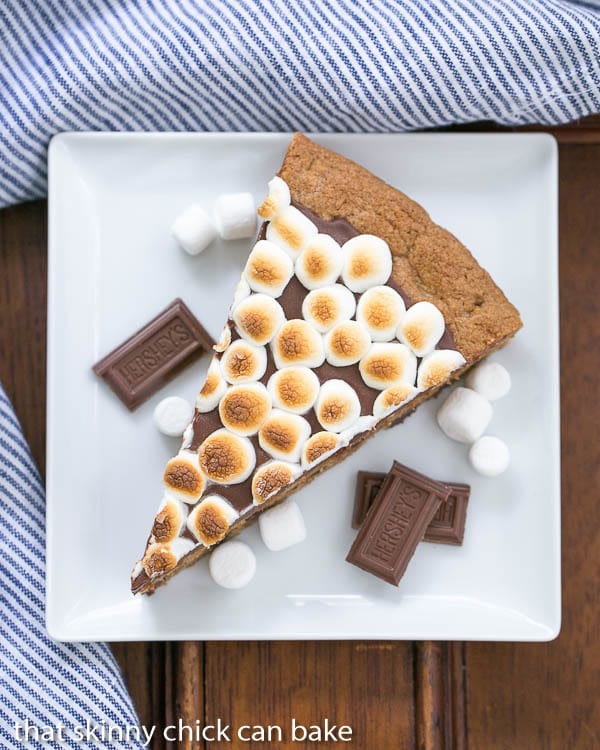 S'mores Cookie Cake wedge on a square white plate with chocolate bar squares and mini marshmallow garnishes
