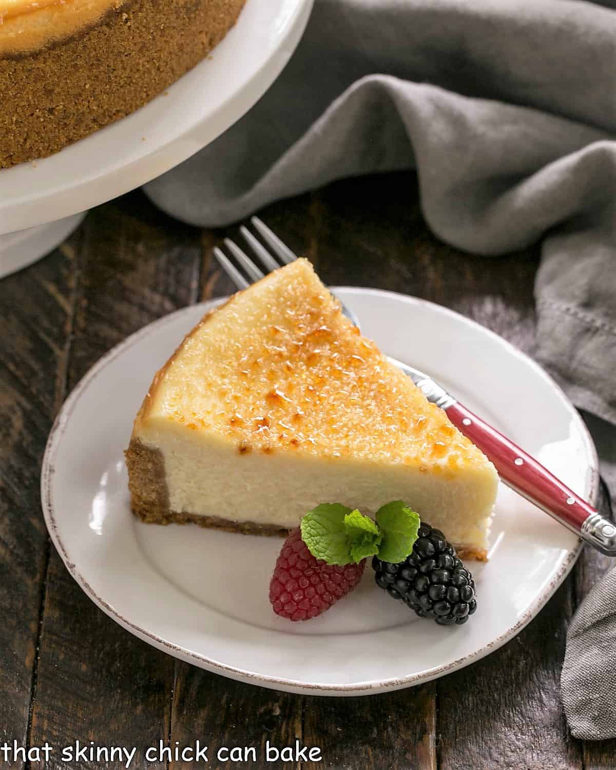 Slice of creme brulee cheesecake on a white dessert plate with a red handle fork, two berries and a sprig of mint.