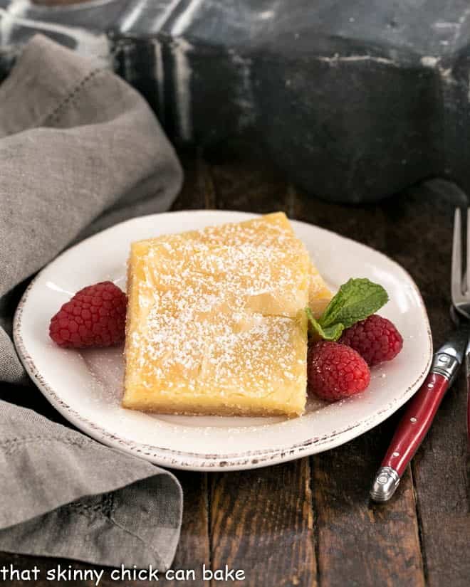 Two gooey butter bars on a round white plate with fresh raspberries and mint.