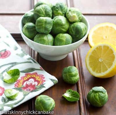 Brussels Sprouts in a small white bowl next to sliced lemons on a wooden table.