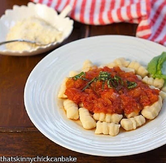 Ricotta Gnocchi topped with marinara and fresh basil on a white dinner plate with a small bowl of Parmesan in the background.