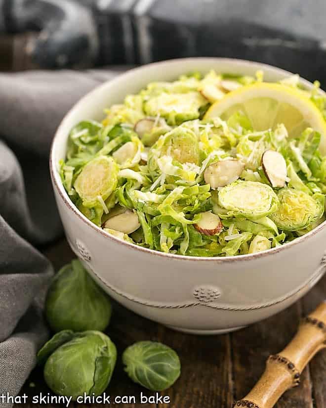 Shaved Brussels Sprouts Salad in a white salad bowl with a lemon slice and a bamboo handled fork.