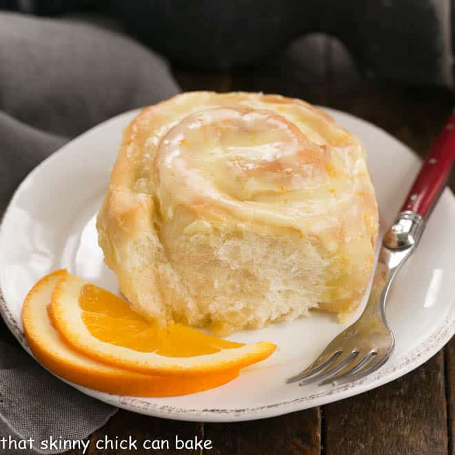 Orange Rolls - a delicious twist on cinnamon rolls filled and iced with orange frosting! Orange Roll on a round white plate with two orange slices and a red handled fork.