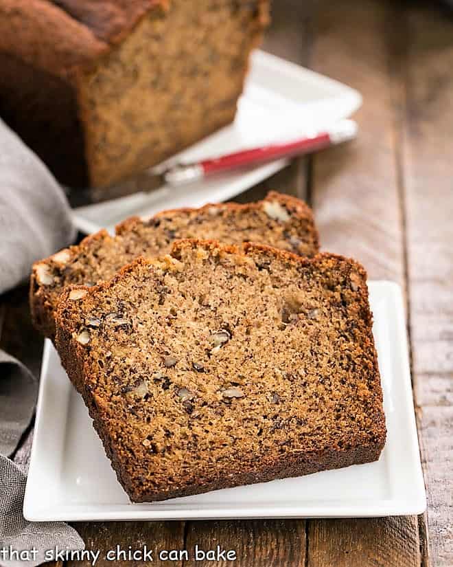 2 slices of Best Banana Bread on a square white plate with the rest of the loaf in the background,