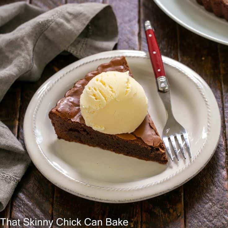 A slice of brownie tart topped with vanilla ice cream on a white dessert plate with a red handle fork