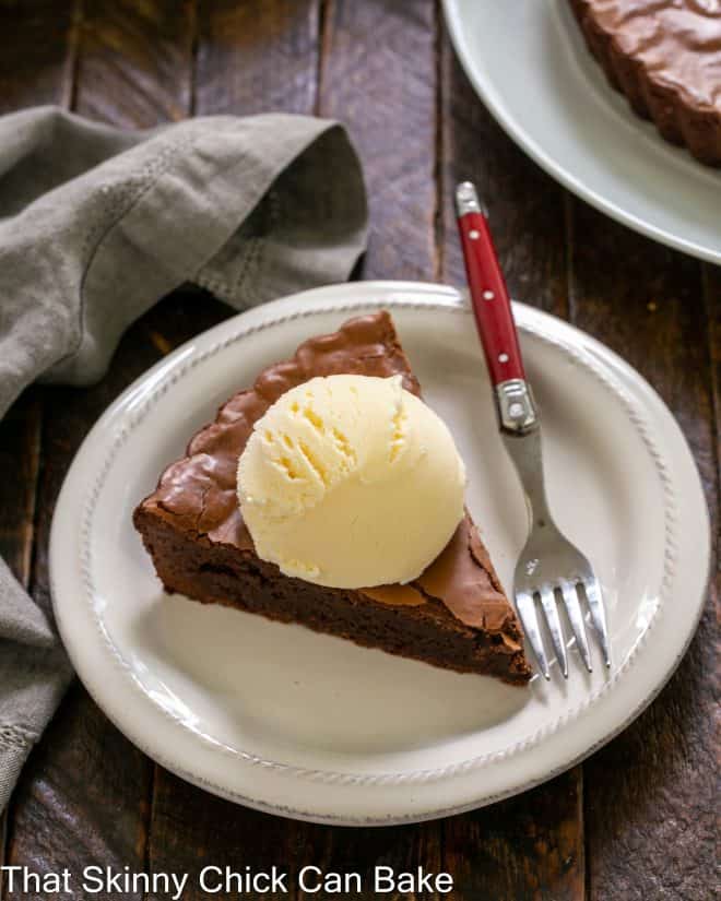 A slice of brownie tart topped with a scoop of vanilla ice cream on a round white plate with a red handled fork.