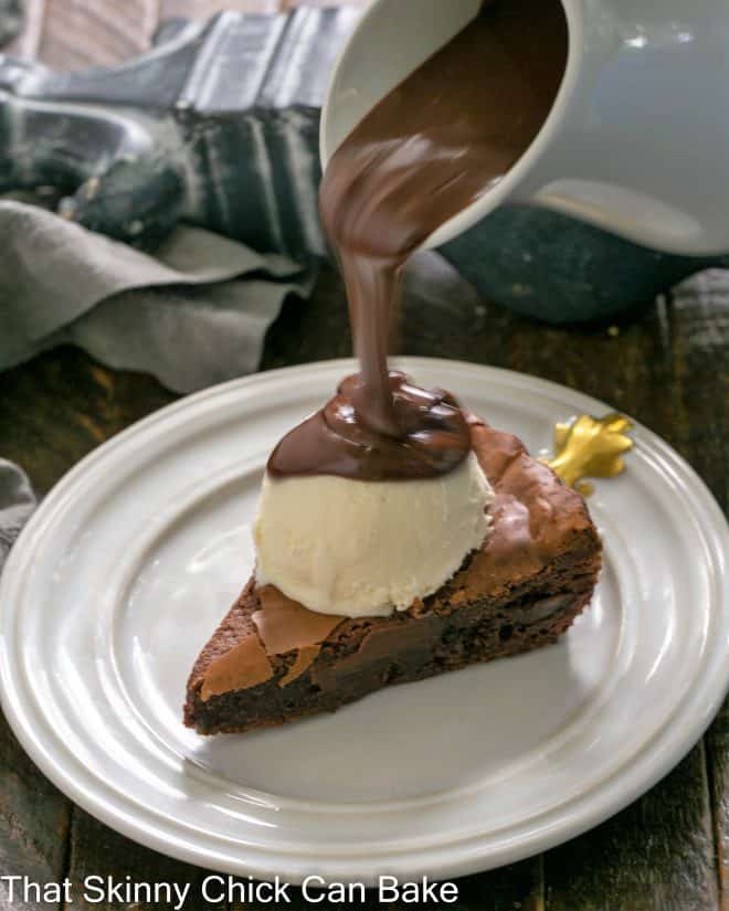 Slice of brownie tart topped with a scoop of vanilla ice cream with chocolate sauce pouring out from a white ceramic pitcher.