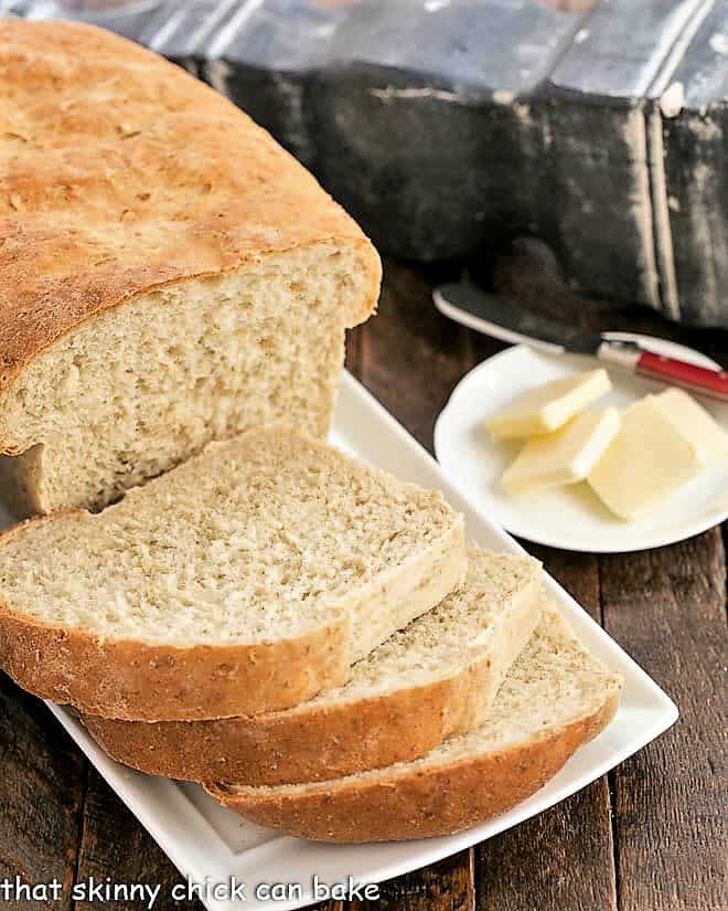 Partially sliced loaf of oatmeal bread on a white tray with a plate of butter pats in the background.