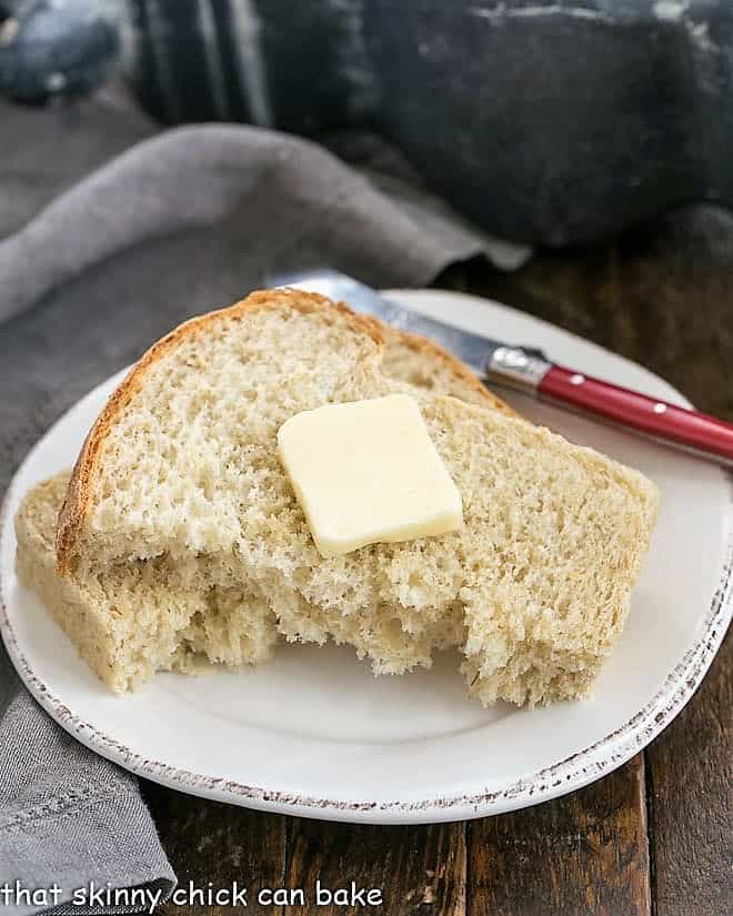 Slice of oatmeal bread torn in half and topped with butter on a round white plate with a red handled butter knife.