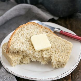 Slice of oatmeal bread torn in half and topped with butter on a round white plate with a red handled butter knife