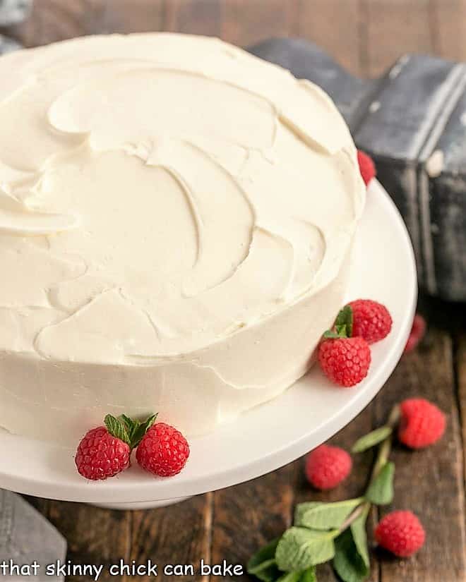 Close up of vanilla cae on a white cake stand wih raspberries and mint garnishes.