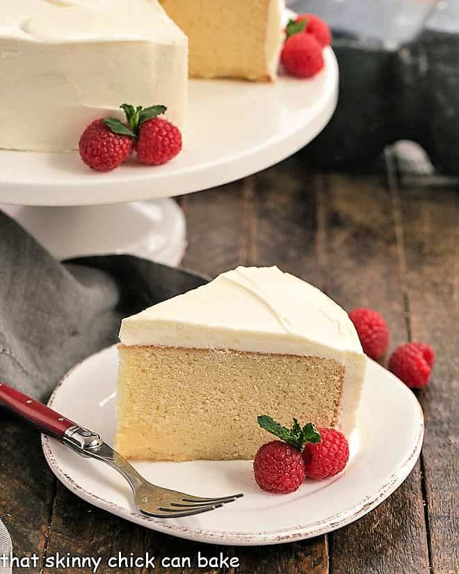 Vanilla Cake with White Chocolate Buttercream on a white cake stand with one slice in the foreground.