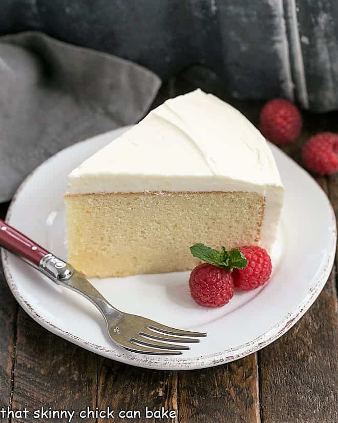 A slice of this white cake recipe on a white dessert plate with a red handled fork and raspberry garnish.