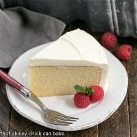 Slice of vanilla cake on a round dessert plate with two raspberries and a red handled fork