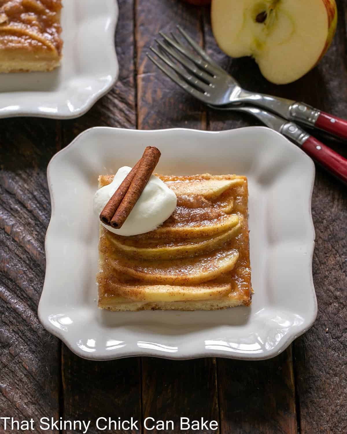 slice of apple kuchen on a square white plate with whipped cream and a cinnamon stick garnish.