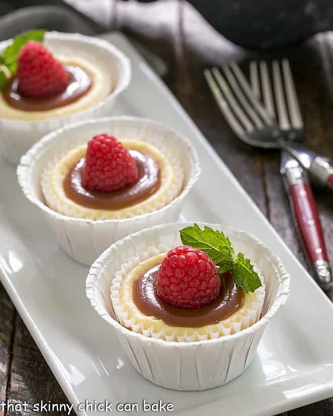 Caramel Topped Mini Cheesecakes on a white rectangular tray garnished with raspberries.