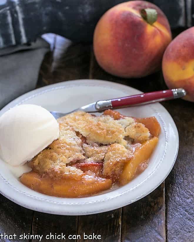 Easy peach cobbler on a white plate with scoop of vanilla ice cream with two fresh peaches in the background.