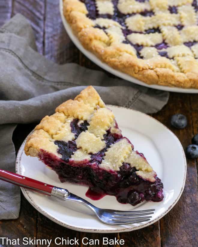 Slice of blueberry pie on a white plate with a red handle fork in front of the remaining pie