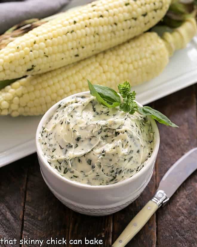 A bowl of basil butter in front of a platter of corn.