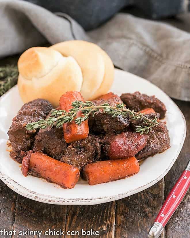 Beef stew with carrots and a sprig of thyme with a dinner roll on a white plate