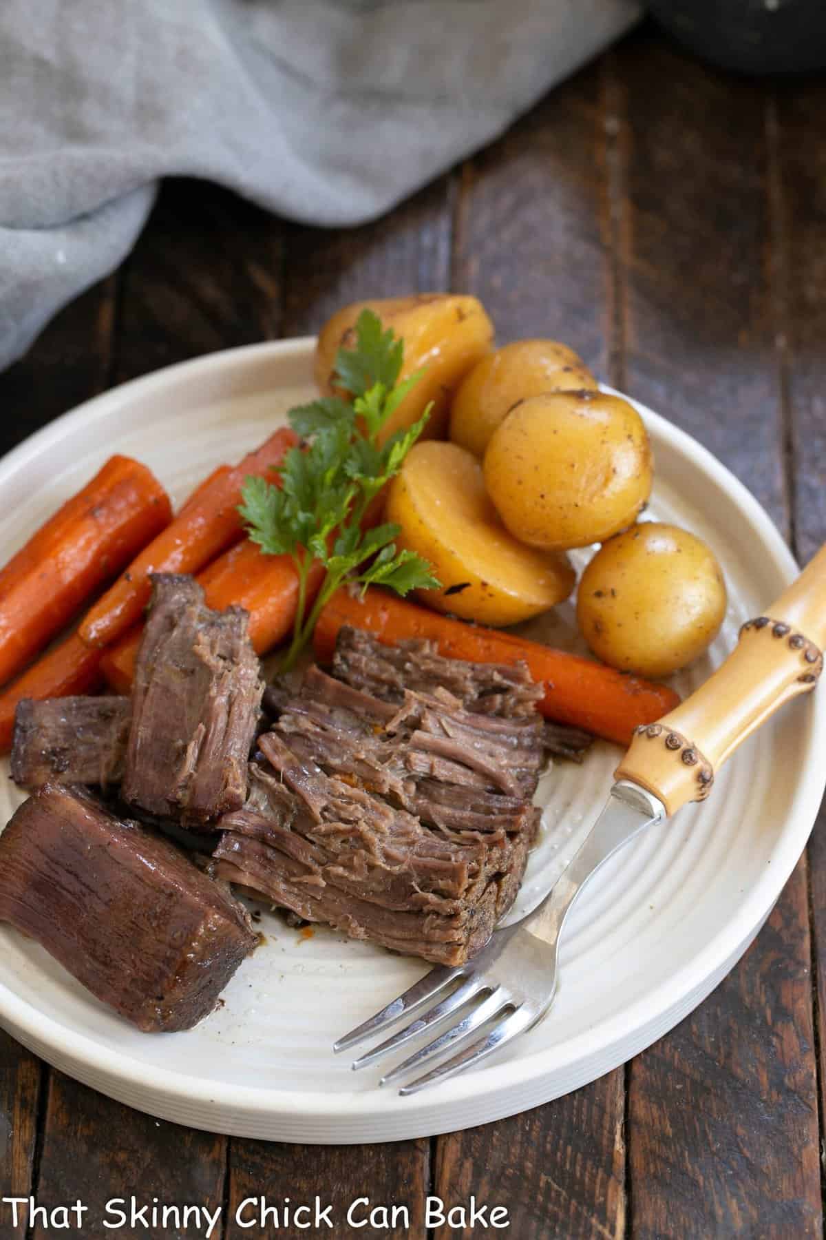 Beef pot roast on a white dinner plate with carrots potatoes and a bamboo handle fork.