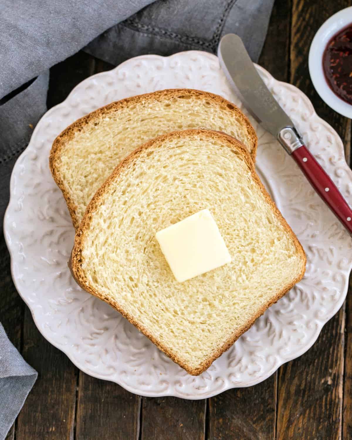 Two slices of a potato bread recipe with a pat of butter and a red-handled knife on a decorative wite plate.