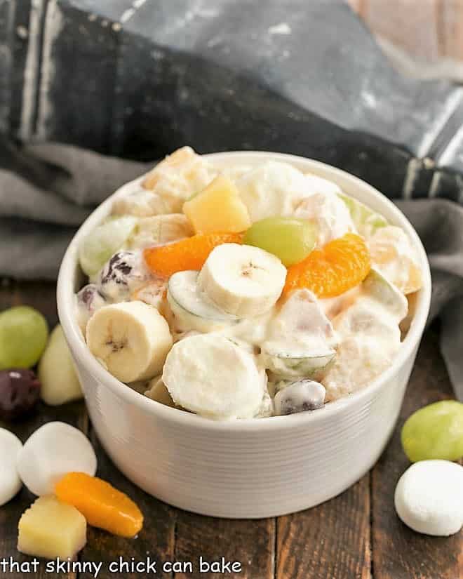 Holiday Fruit Salad with Marshmallows in a white ceramic bowl surrounded by fruit and marshmallows