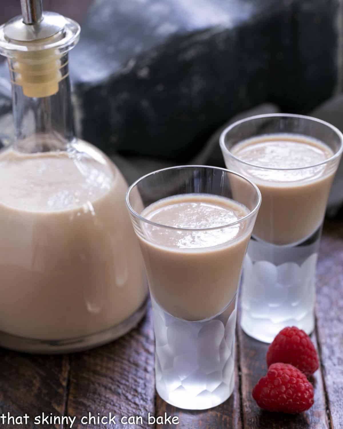 Irish cream recipe in a carafe and two shot glasses of Baileys with some fresh raspberries.